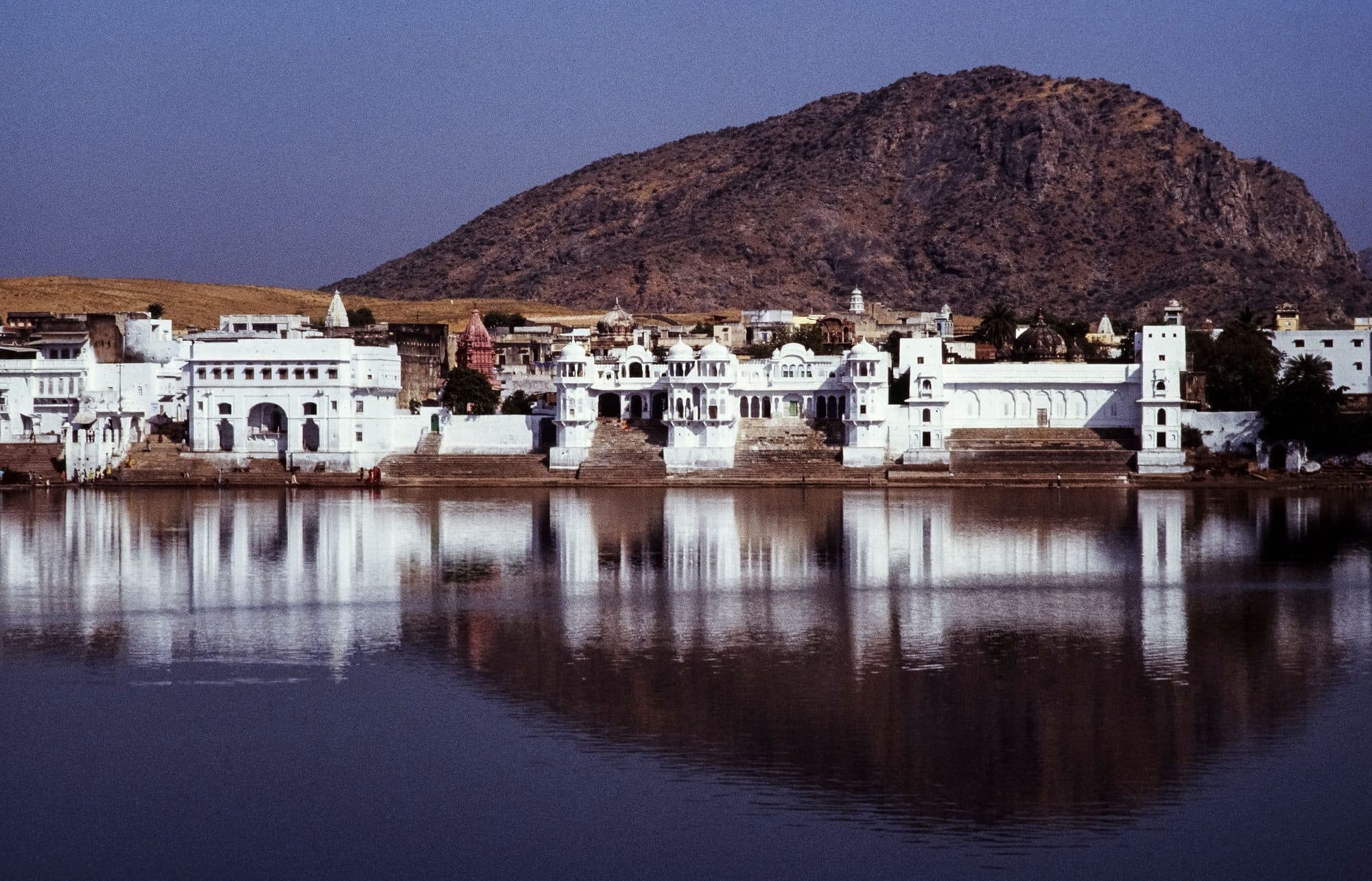 Whitewashed ghats and temples of Pushkar Lake reflected in still water, with a rocky hill behind